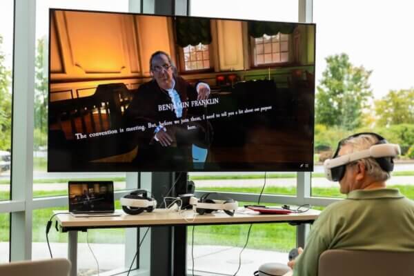 An individual wears a virtual reality headset as a TV displays a virtual avatar of Benjamin Franklin in Independence Hall.