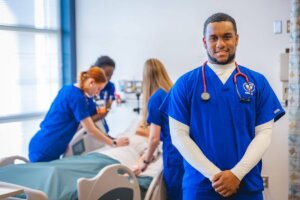 Shenandoah University nursing students, wearing blue scrubs, gathered around a manikin (simulated patient) in a bed.