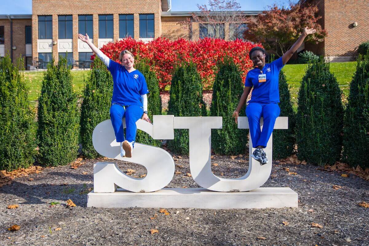 Two Shenandoah nursing students, in blue scrubs, sit atop the SU statue in the Shenandoah University Quad in the autumn.