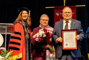 Wilbur and Clare Dove stand on the commencement stage with Shenandoah University President Tracy Fitzsimmons.