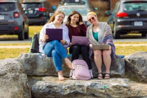 Three Shenandoah University students use their Macbook computers outdoors.