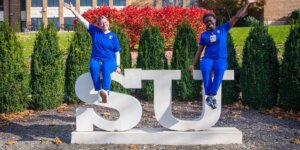 Two Shenandoah nursing students, in blue scrubs, sit atop the SU statue in the Shenandoah University Quad in the autumn.