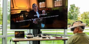 An individual wears a virtual reality headset as a TV displays a virtual avatar of Benjamin Franklin in Independence Hall.